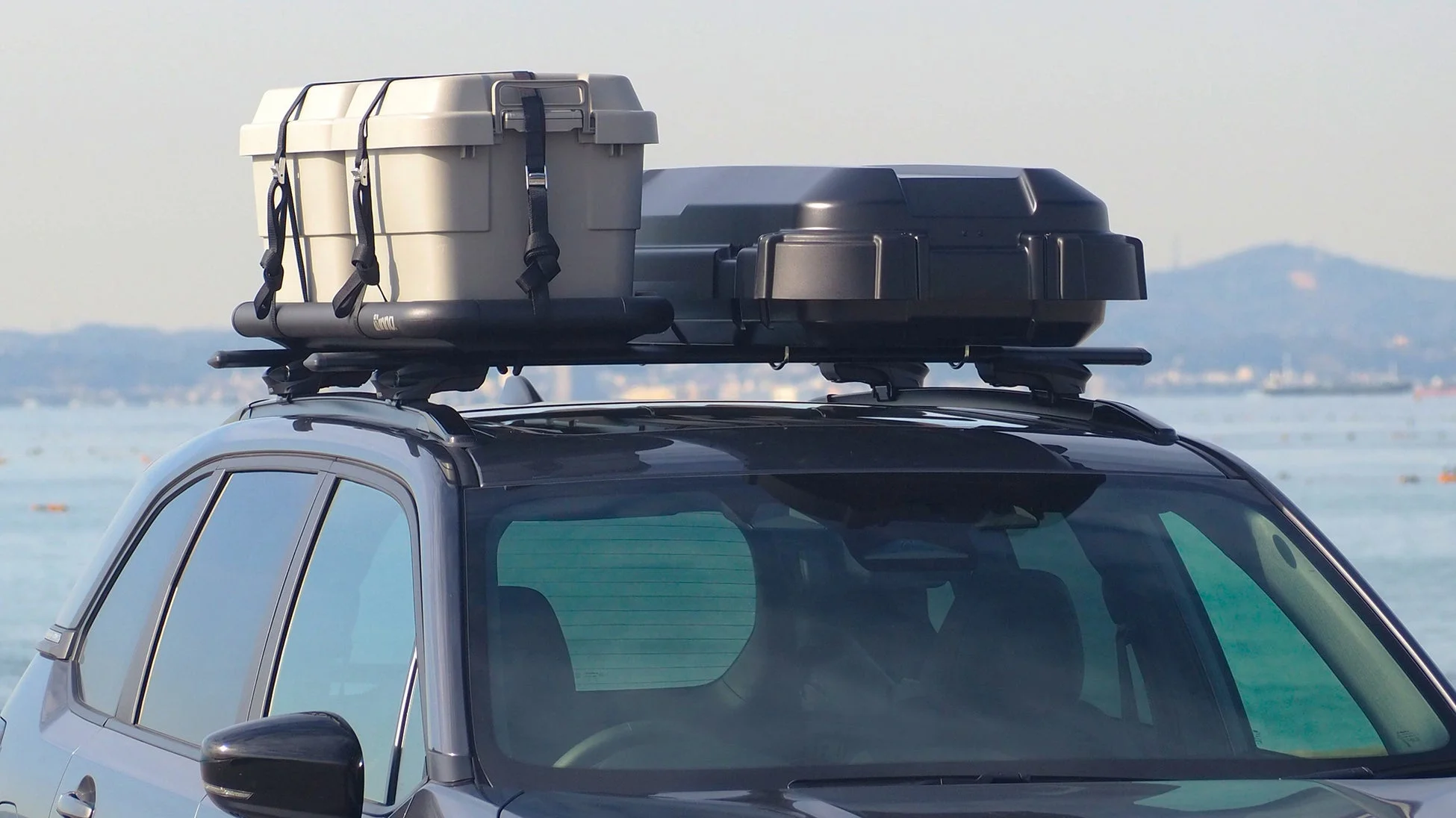 海を背景に、ルーフラックにキャンプ用品のような収納ボックスを積んだSUVの側面が写っています。アウトドアやレジャーに出かける準備が整っている様子です。