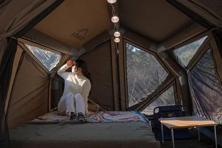 テントの中で女性がベッドに座り、窓から差し込む朝日を眺めている様子
