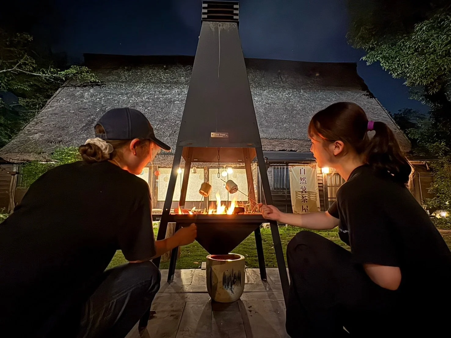 夜、古民家風の建物の前で、2人の女性が焚き火を囲んでマシュマロを焼いている様子です