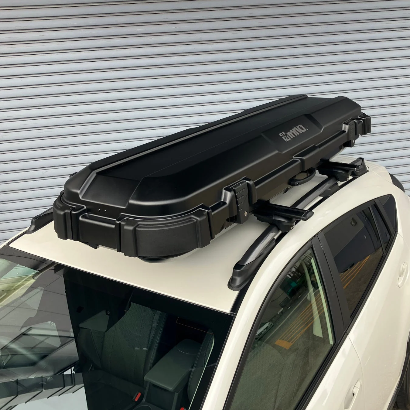 白い車の屋根に黒いルーフボックスが取り付けられています。ルーフラックにしっかりと固定されており、アウトドアや旅行での荷物積載に便利な様子が伺えます。背景はシャッターです。