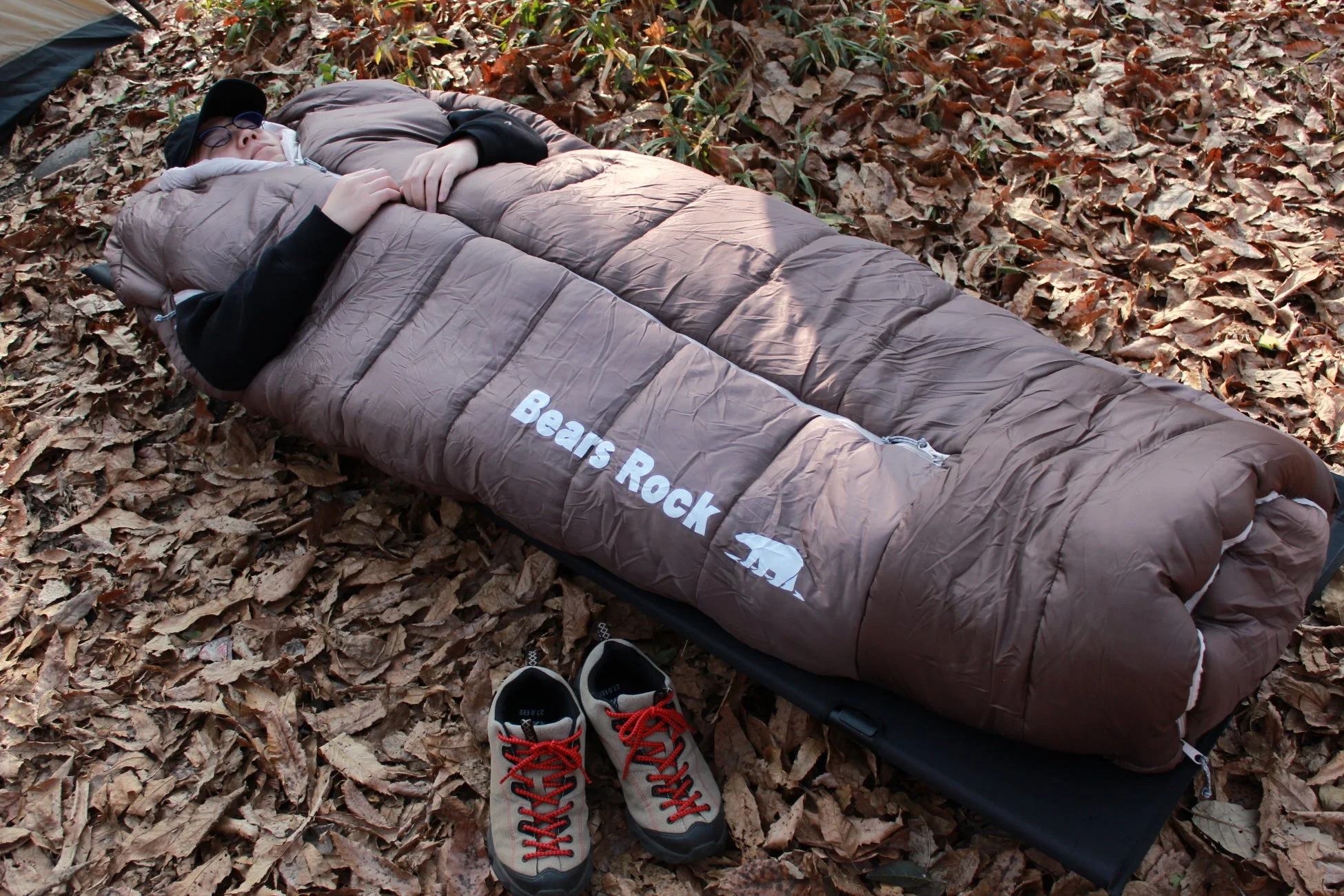 枯れ葉の地面の上で、茶色の寝袋にくるまって寝ている人が写っています。傍らにはハイキングシューズが置かれており、アウトドアでの休息やキャンプの様子がうかがえます。