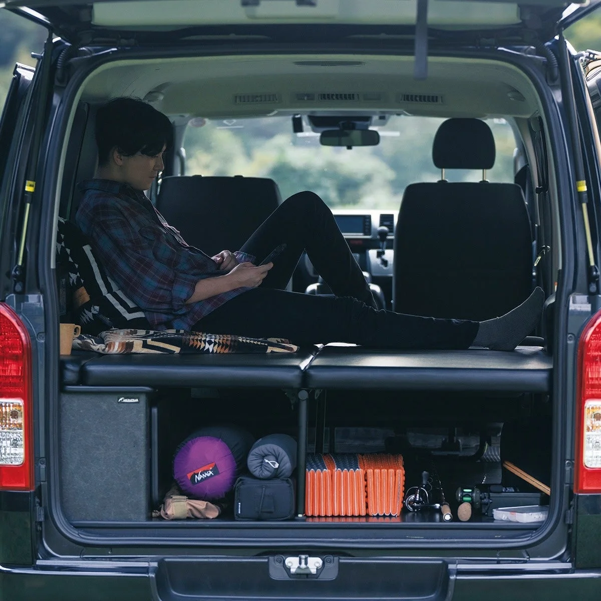 車中泊仕様のバンの後部で、男性がスマートフォンを操作しながらリラックスしている。