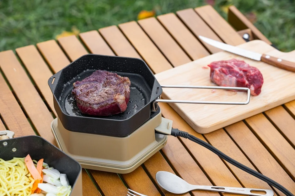 屋外の木製テーブルで、ポータブルコンロを使ってステーキを焼いている様子