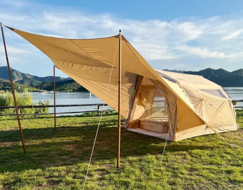 湖畔の広々としたキャンプ場で、ベージュ色の大型テントとタープが設置されています。青空の下、遠くには山々が見え、自然を満喫できる穏やかなアウトドアシーンです。