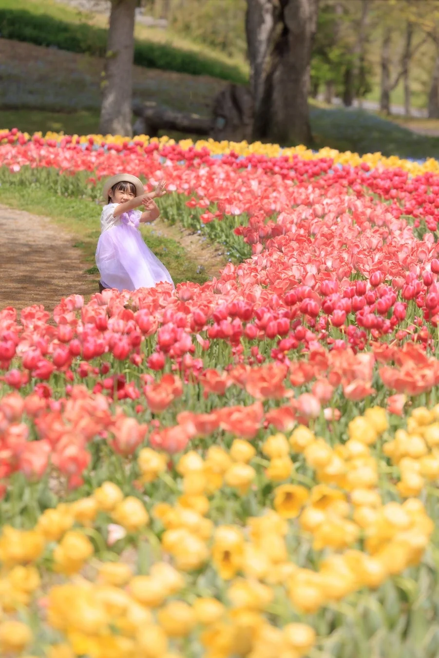 色とりどりのチューリップが咲き誇る花畑で、麦わら帽子をかぶった女の子が楽しそうにポーズをとっている春の風景