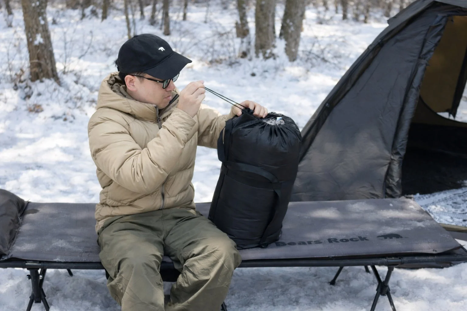 雪が積もった森の中で、男性がコットに座り、寝袋の準備をしている様子