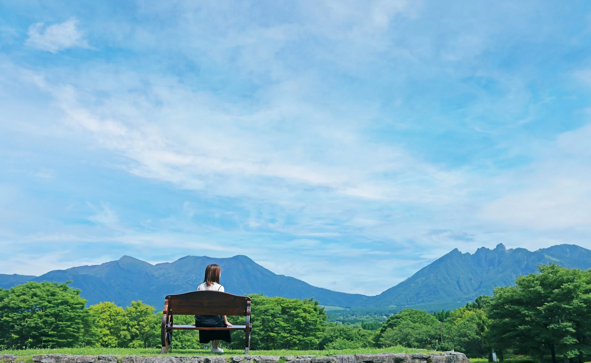 広大な自然の中で、一人の女性がベンチに座り、遠くの山々と青い空、そして緑豊かな景色を眺めている後ろ姿の画像です。穏やかで開放的な風景が広がっています。