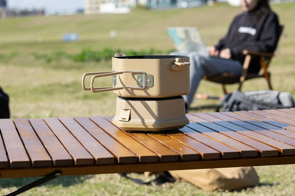キャンプ用テーブルに置かれたクッカーが手前にあり、奥では人物が椅子に座ってくつろいでいるアウトドアシーン