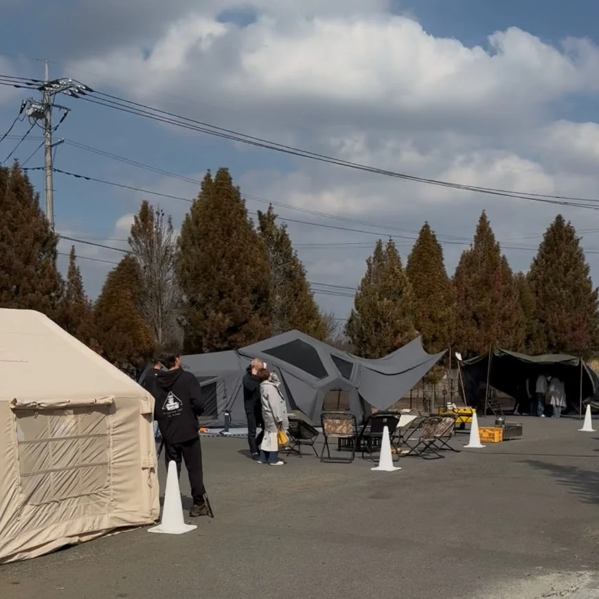 青空の下、人々が複数のテントやキャンプ用の椅子を設営している様子が写っています。アウトドアイベントやキャンプ場の準備風景に見え、木々が背景に広がっています。