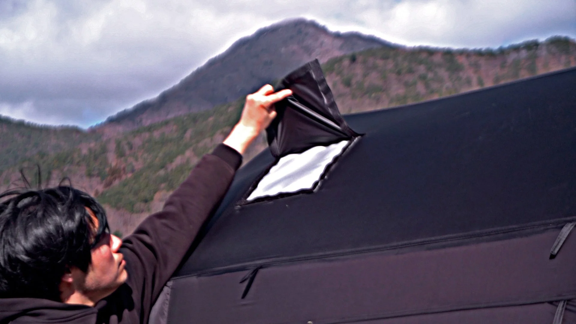 山を背景に、人物が黒いテントの窓の蓋を開けている