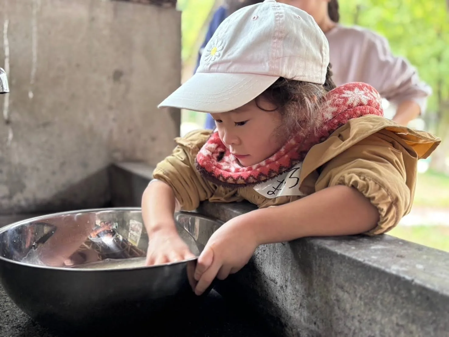 白い帽子をかぶった子供が、屋外のシンクで大きな金属製のボウルに入った水に手を入れ、何かに夢中になっている様子