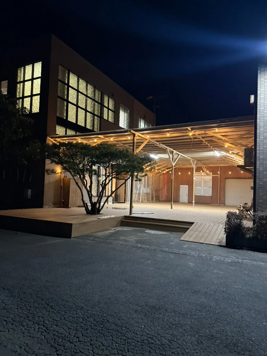 夜の屋外スペースを捉えた画像です。明るく照らされた建物の窓、ウッドデッキ、一本の木、そして電球で飾られた屋根付きのエリアが見えます。静かで落ち着いた雰囲気です。