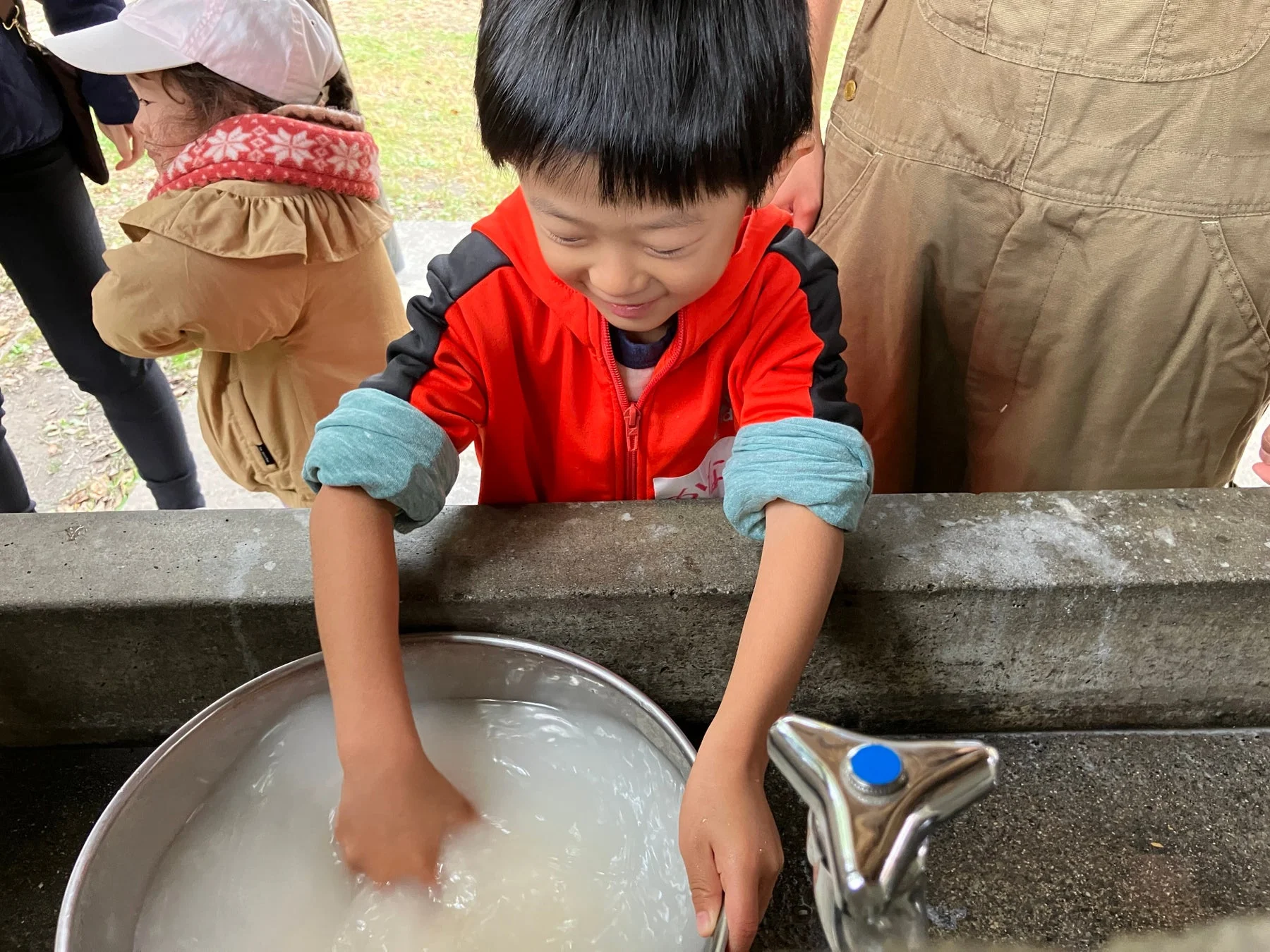 笑顔の子供が、屋外で大きな金属製のボウルに入った白濁した水をかき混ぜ、何かを洗っている様子です