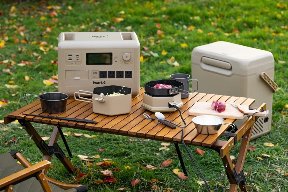 秋の景色の中で、ポータブル電源やクーラーボックスなどのキャンプ用品が並んだ木製テーブルで野外料理をしている様子