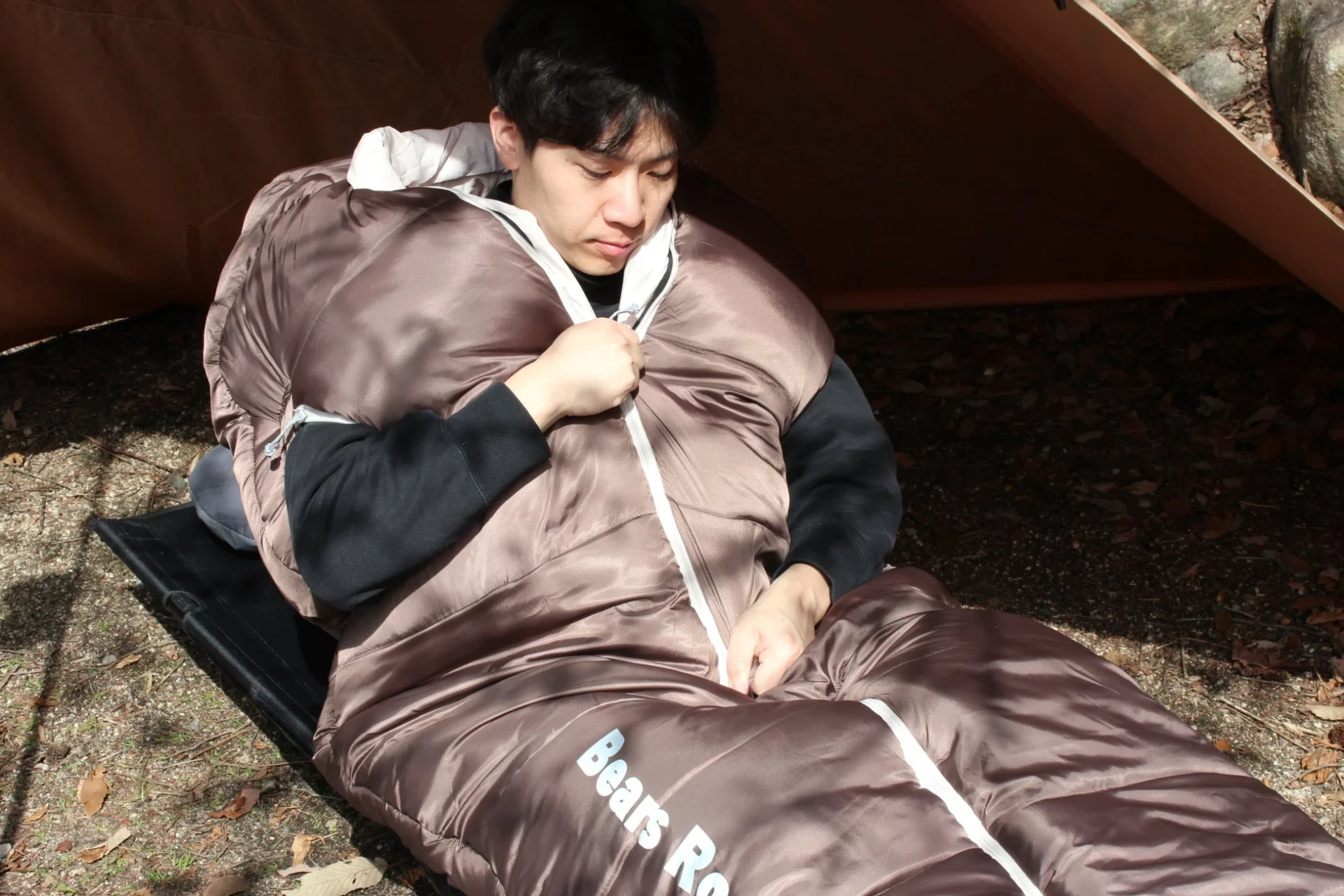 屋外で茶色の寝袋に入り、コットに横たわっている若い男性
