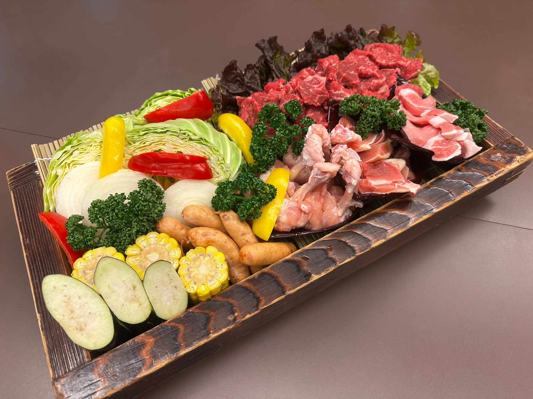 焼肉やBBQ、鍋物用の食材が盛り付けられた豪華なプレート。牛肉、豚肉、鶏肉、ソーセージに加え、キャベツ、パプリカ、玉ねぎ、ナス、トウモロコシなど彩り豊かな野菜が豊富に揃っています。