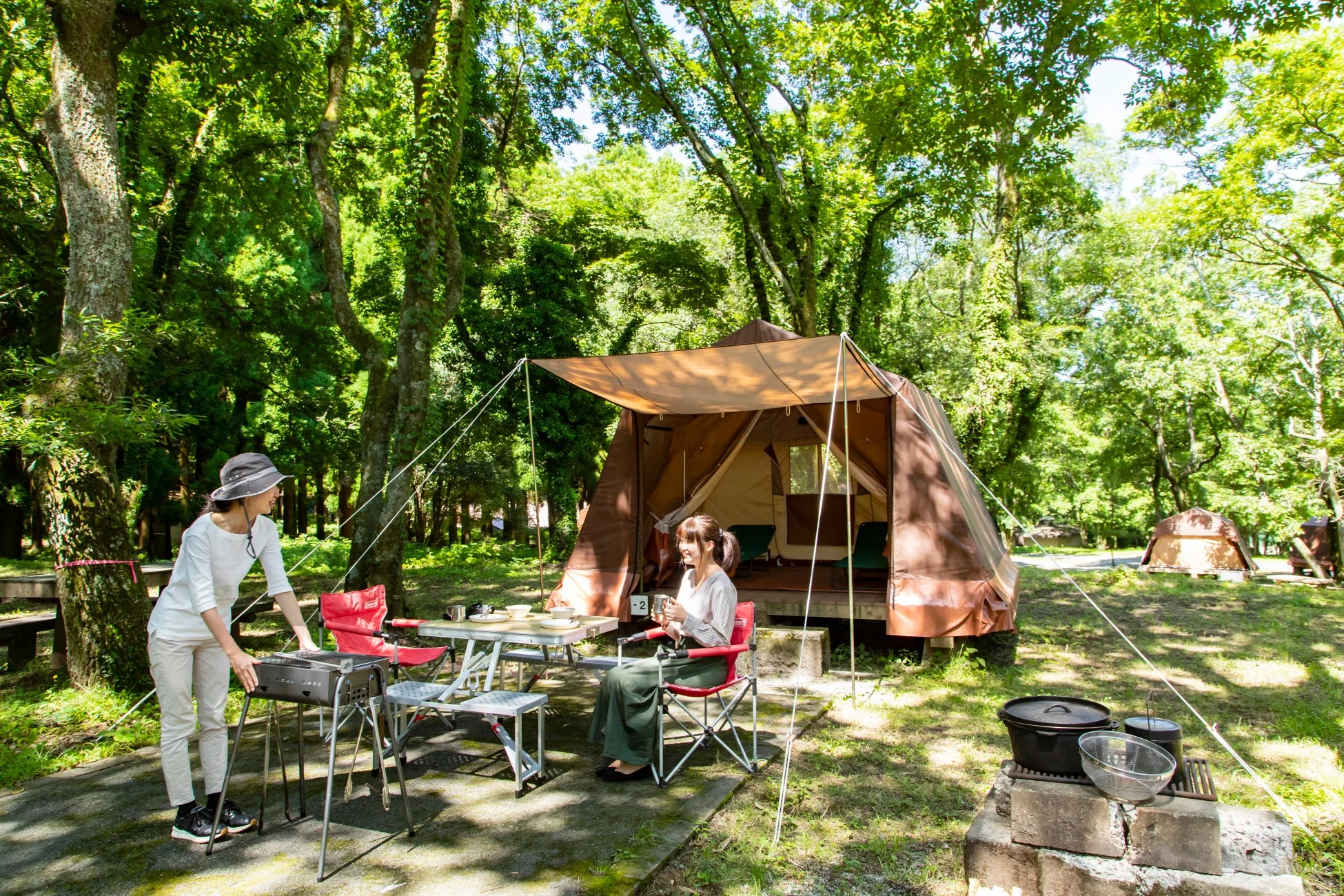 緑豊かな森林の中で、2人の女性がキャンプを楽しんでいます。一人はバーベキューの準備をし、もう一人はテントのそばでくつろいでおり、穏やかな休日の雰囲気が伝わってきます。