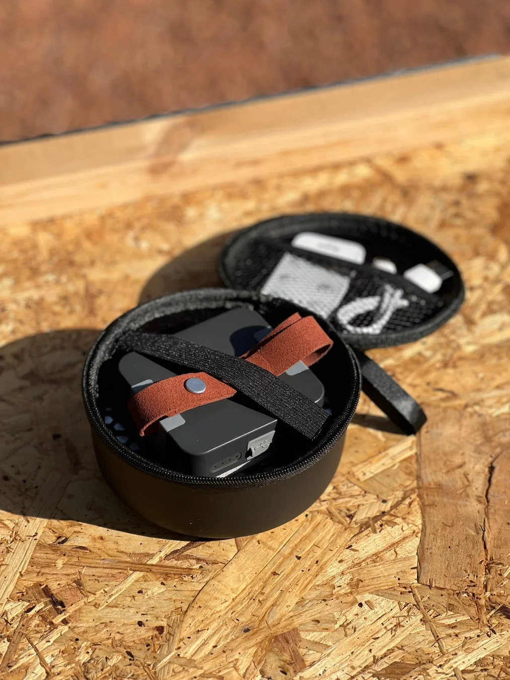 木製の表面に置かれた開いた黒いケースに、茶色のストラップが付いた小型の電子機器と、蓋のメメッシュポケットにケーブルなどのアクセサリーが収納されている様子