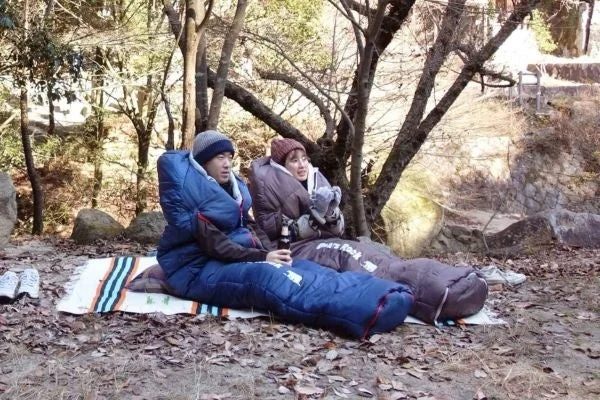 木々に囲まれた自然の中で、男女が寝袋のような防寒着を着て座っている