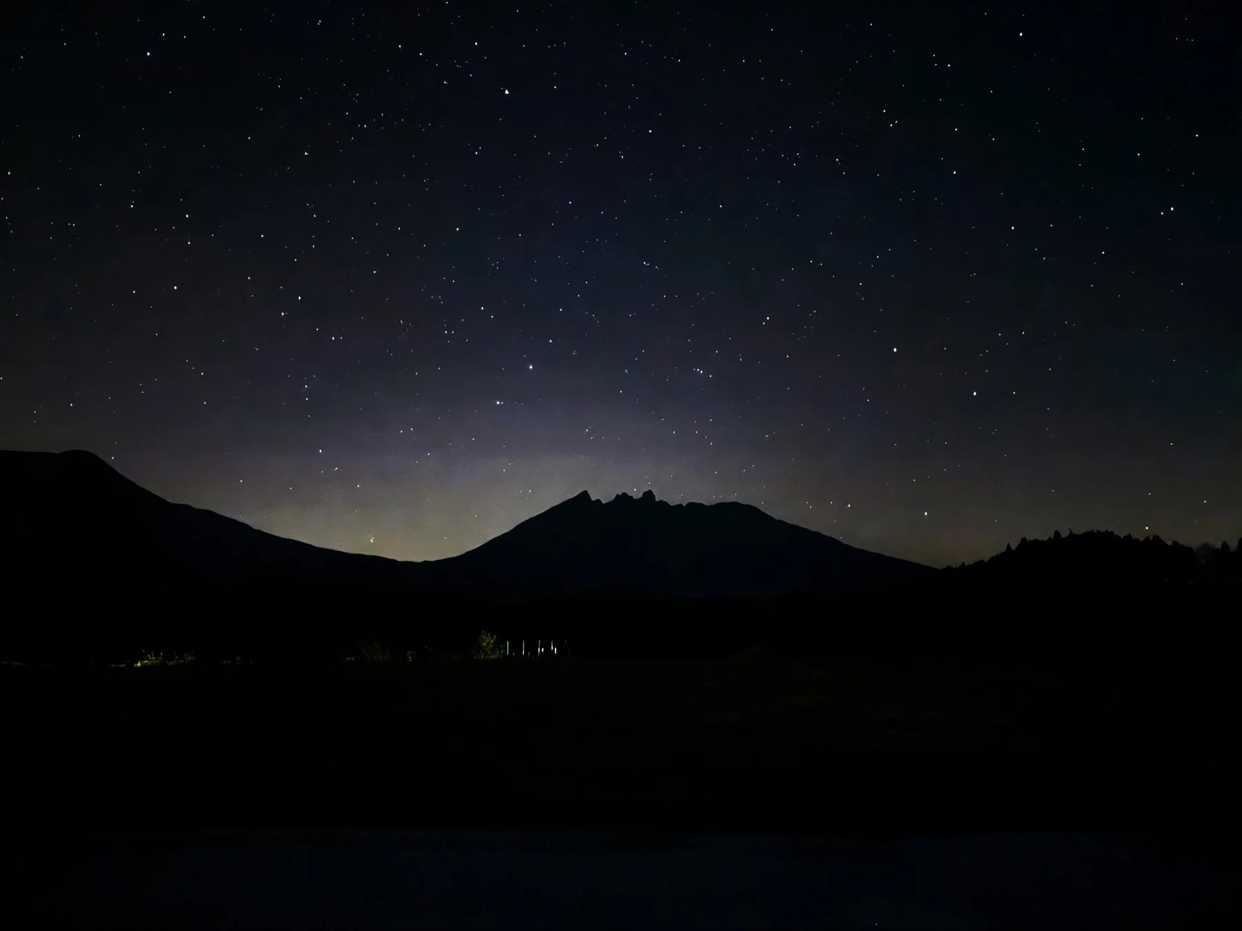 満天の星が輝く夜空と、その下に広がる山々のシルエットが印象的な夜景です。遠くには微かな光が見え、静かで神秘的な雰囲気を醸し出しています。