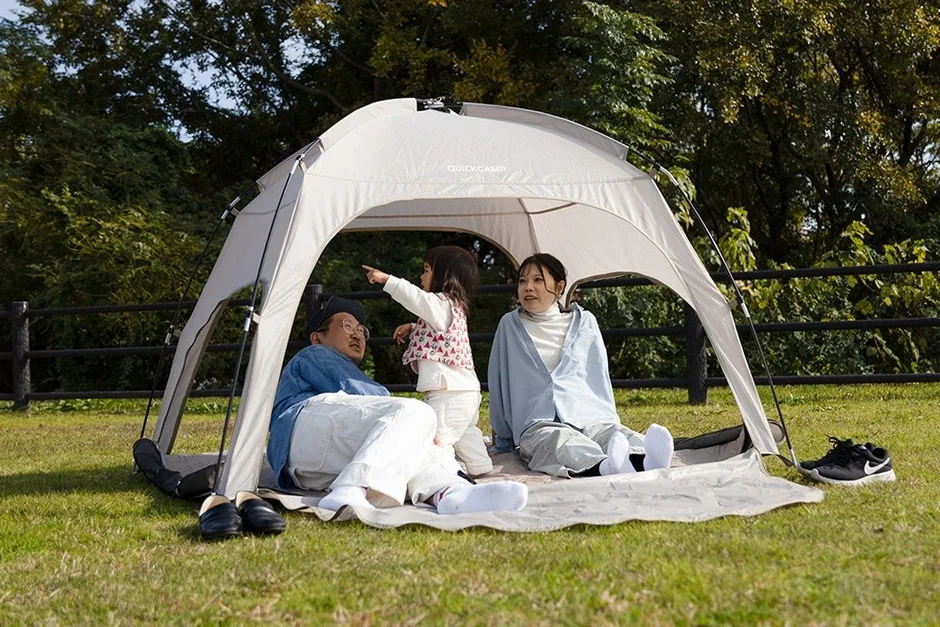 晴れた日に家族3人が公園の芝生に設置されたテントの下でくつろいでいます