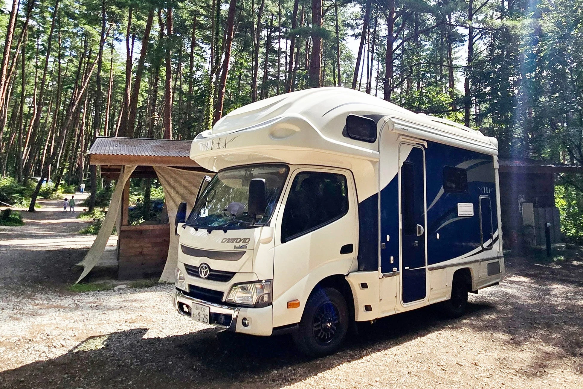 晴れた日に森の中のキャンプサイトに駐車されている、白と紺色のキャンピングカー