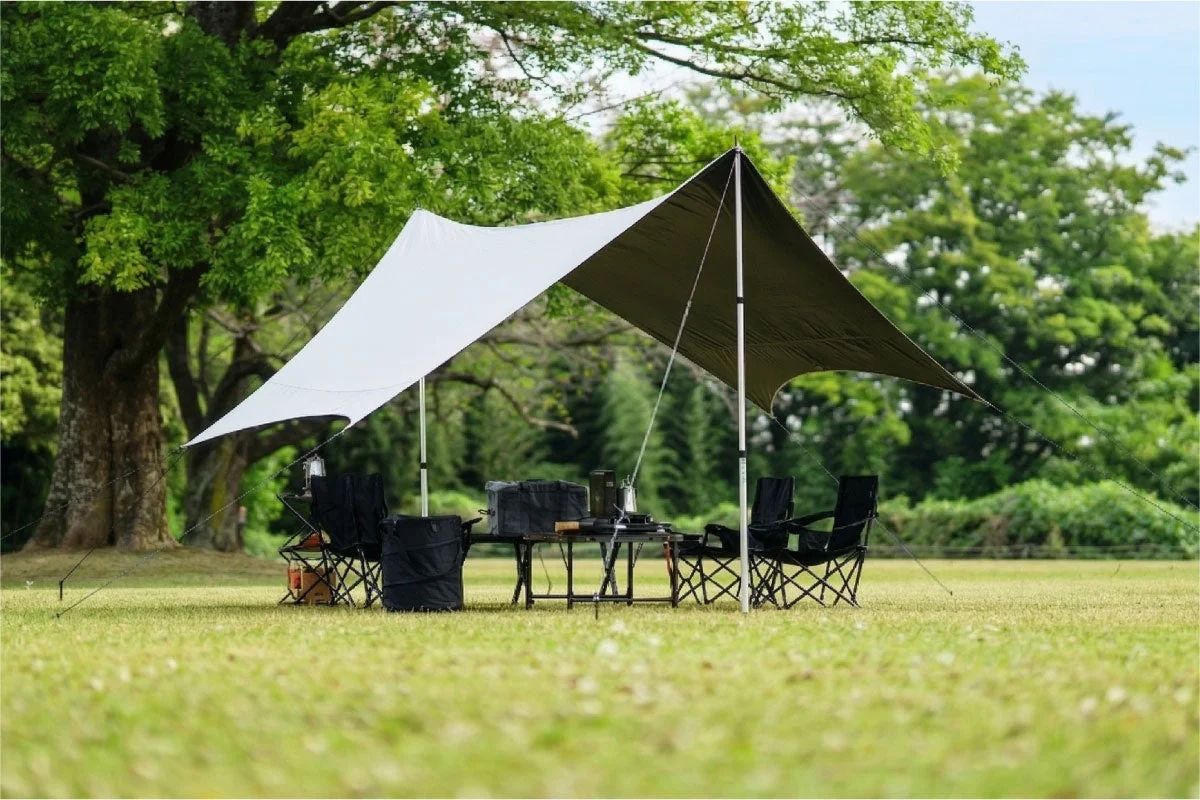 広々とした芝生の広場に、大きな木を背景にしてキャンプ用のタープが張られています。タープの下にはテーブルと複数の折りたたみ椅子が配置されており、アウトドアでの食事や休憩を楽しめる準備が整っている様子です。