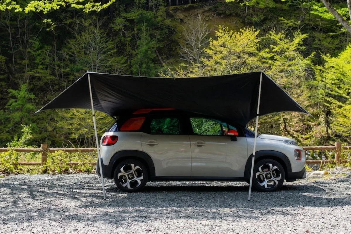 砂利の地面に停められた淡い色のSUVの上に黒いタープが張られ、日差しを避けている様子。豊かな緑の木々と木製の柵が見え、アウトドアやキャンプの雰囲気が漂う一枚です。