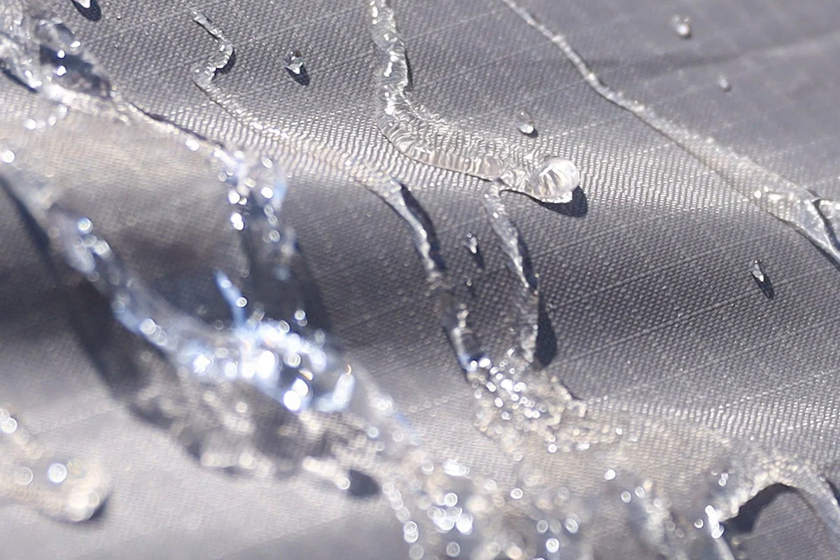 暗い色の撥水性のある生地の表面に、水滴や水の流れがクローズアップで捉えられています。水が玉状になって生地の上を滑り落ちる様子が鮮明に写し出されており、素材の防水・撥水性能を示唆しています。