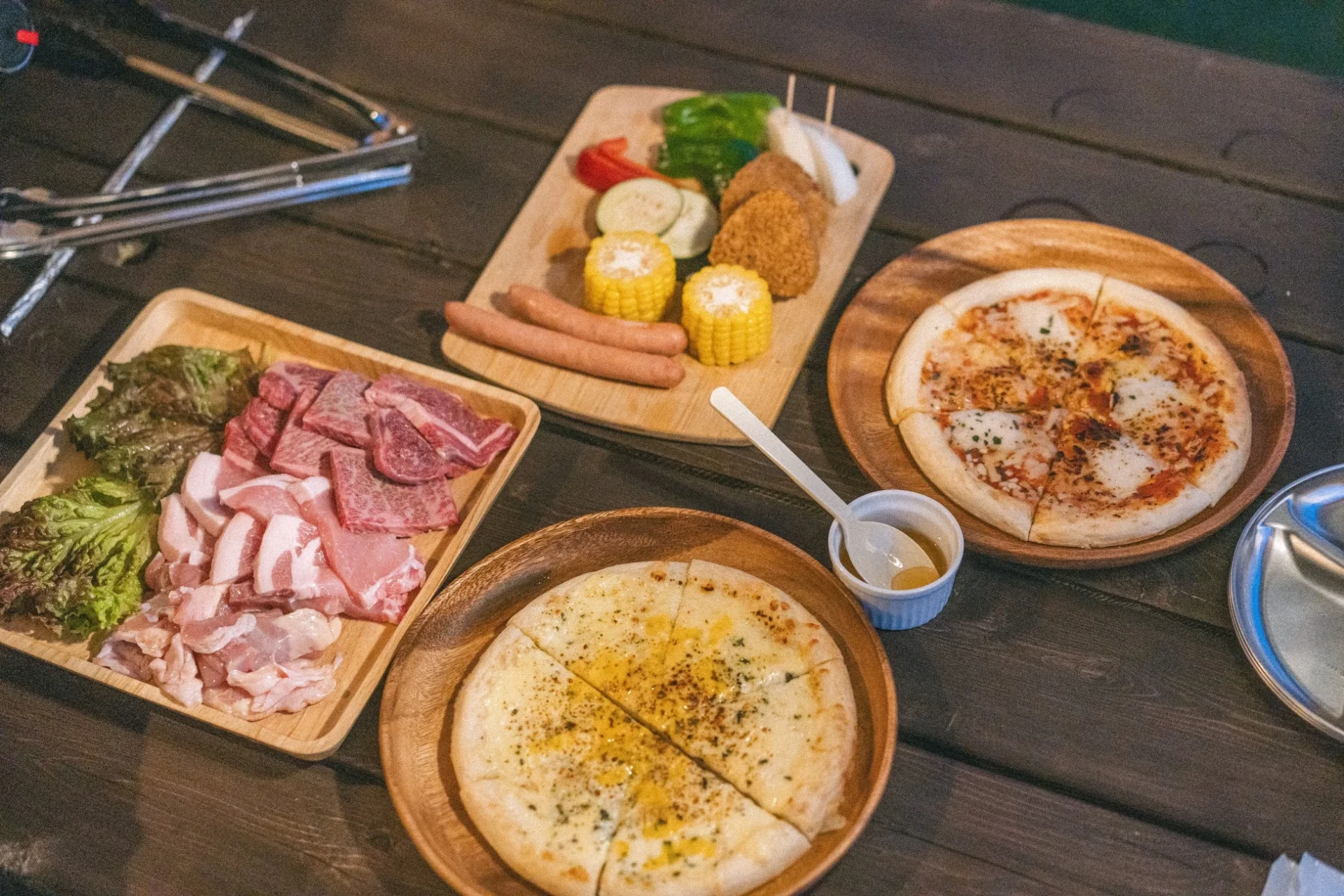 木製のテーブルに、焼肉用の生肉、ソーセージ、野菜、揚げ物、そして2種類のピザが並べられています