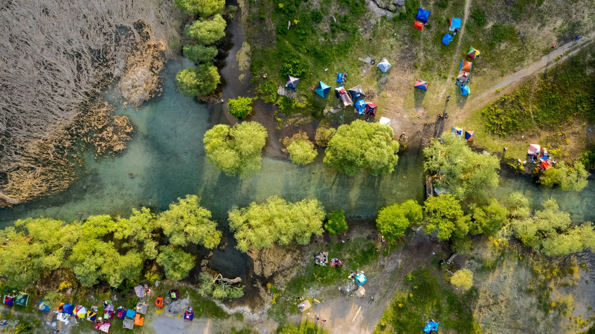 豊かな緑と乾いた葦に囲まれた曲がりくねった川沿いに広がる、賑やかなキャンプ場の空撮写真です。色とりどりのテントが多数張られ、人々がアウトドアを楽しんでいます。