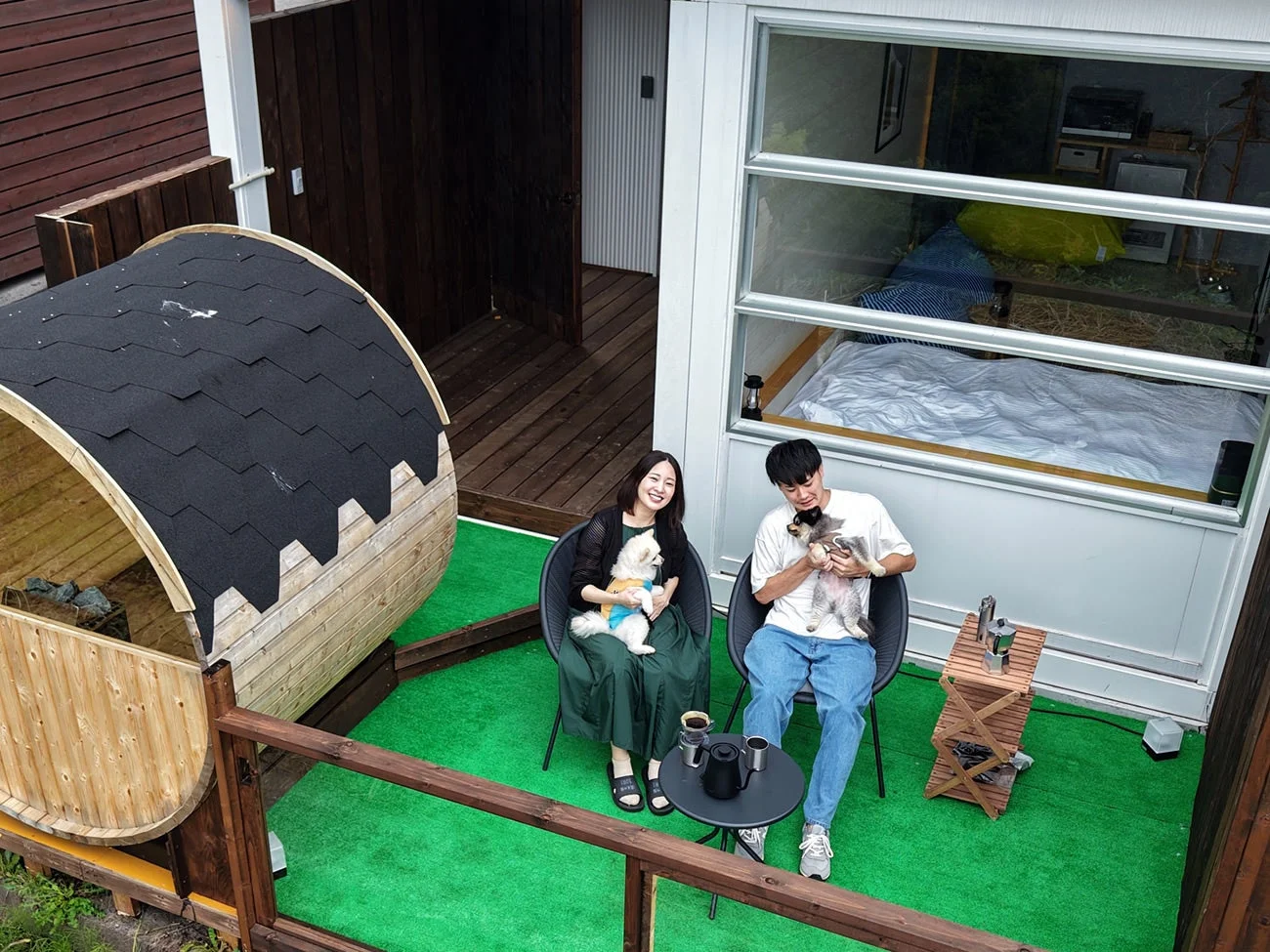 緑の人工芝の上で、若い男女がそれぞれ小型犬を抱き、コーヒーを飲みながらリラックスしている様子