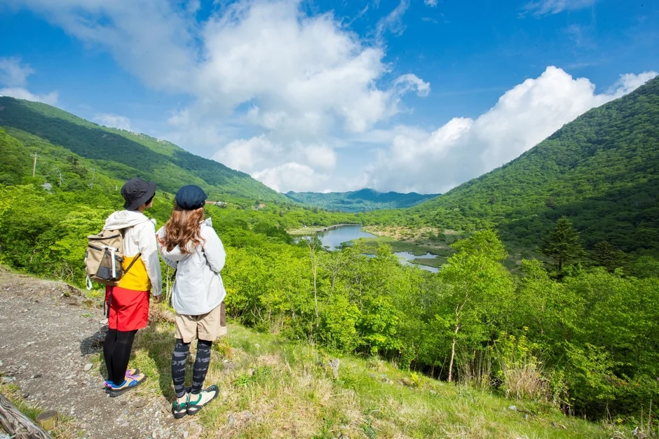 2人のハイカーが、新緑の山々と湖が広がる雄大な自然の中で景色を眺めている様子