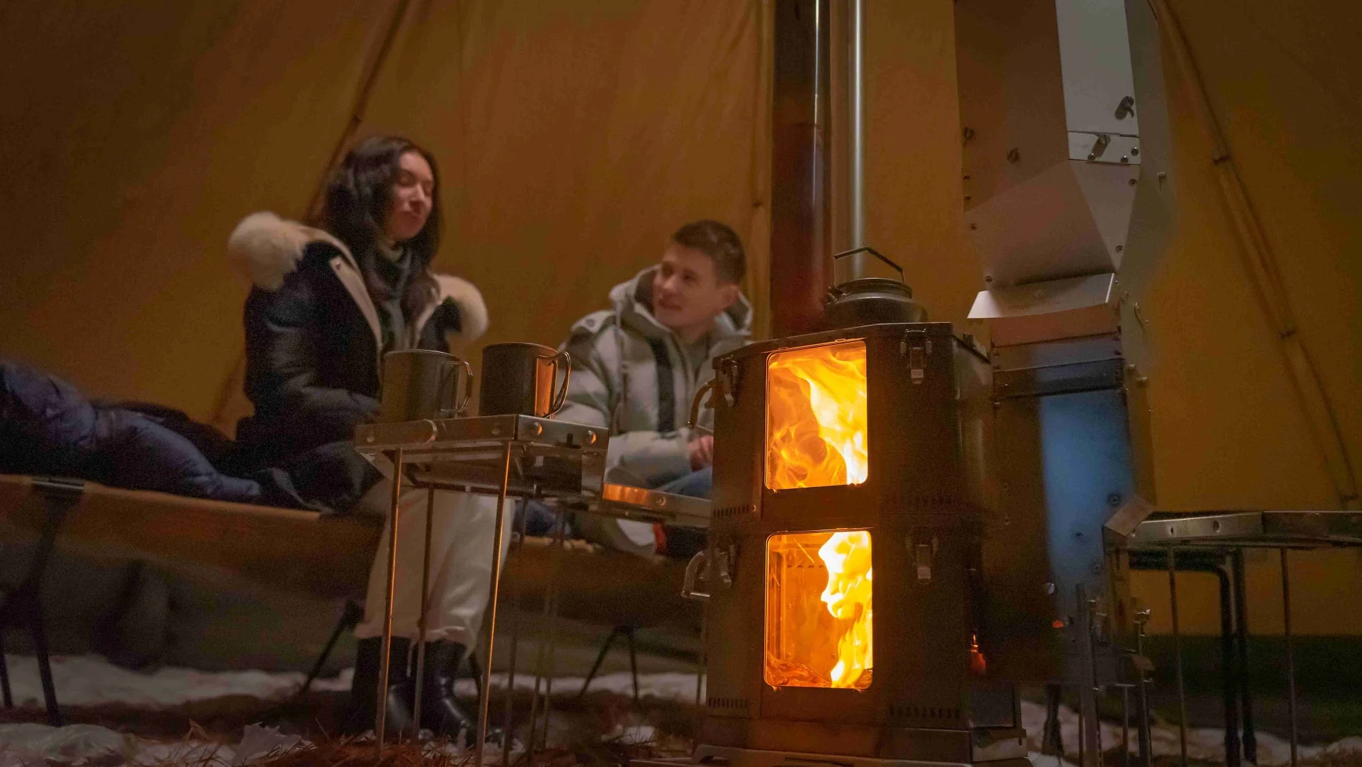 暖かいテントの中で、薪ストーブの炎を囲んで談笑する男女の様子