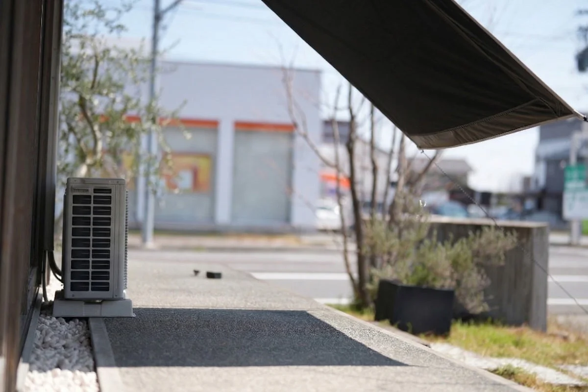 建物の外壁沿いに設置されたエアコンの室外機が手前にあり、上部には日よけの庇が写っています。奥には道路を挟んで別の建物が見え、晴れた日の屋外の風景が広がっています。