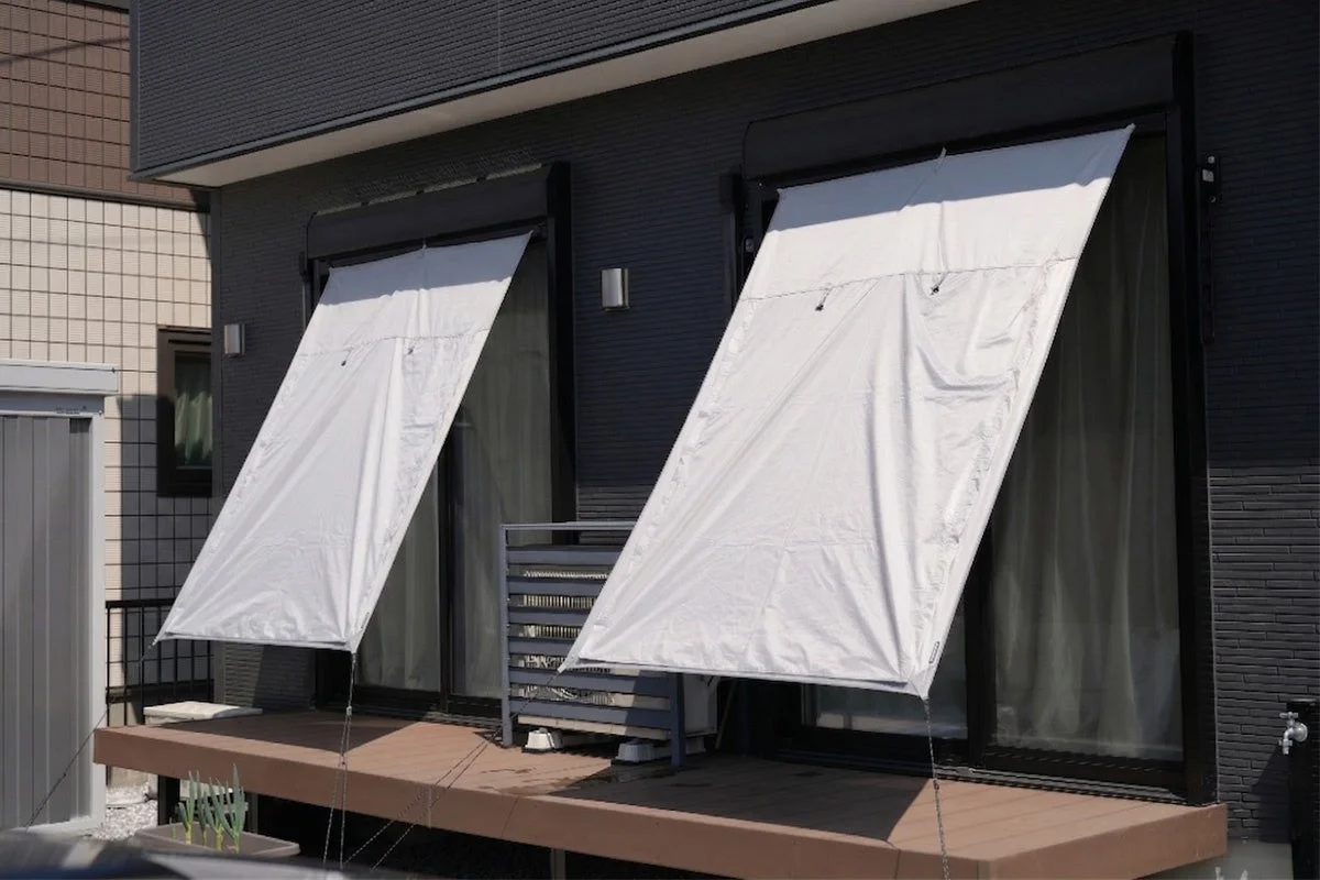 現代的な住宅の窓に、夏の強い日差しを遮るための白い日よけ（シェード）が取り付けられています。手前には木製のデッキがあり、建物の外壁はダークグレーで統一されています。