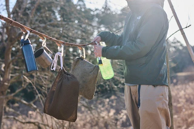 屋外で男性が木の枝に水筒やマグカップ、収納袋などのキャンプギアを吊るして整理している様子