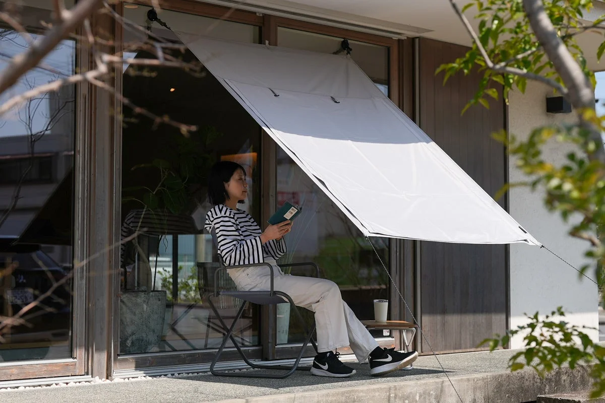 日当たりの良いテラスで、白い日よけの下、女性が椅子に座って読書を楽しむ穏やかな情景。くつろいだ午後のひとときが感じられる。