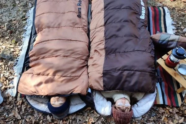 落ち葉が敷き詰められた屋外で、二人が寝袋に入って横たわっている