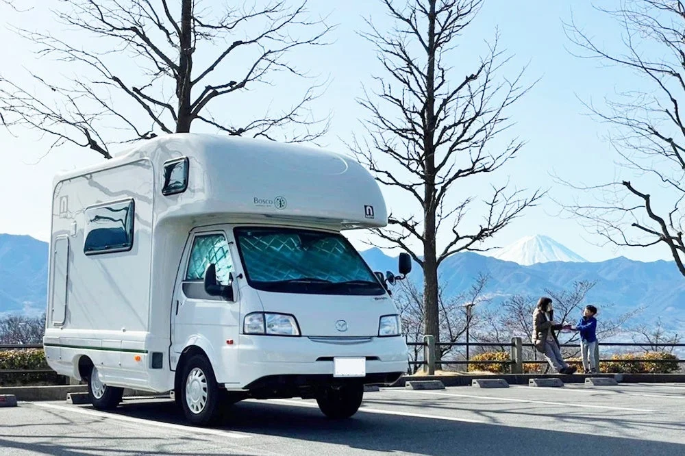 冬の晴れた日に、白いキャンピングカーが駐車場に停まり、背景に雪をいただいた富士山がそびえ立つ風景