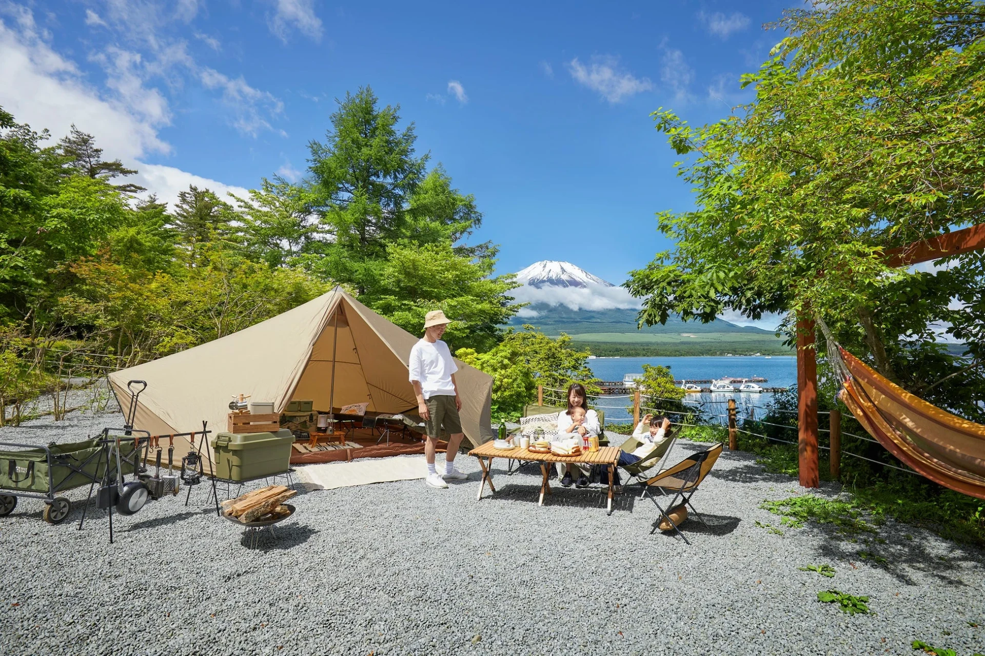 晴れた日、富士山と湖を望むキャンプ場で、家族がグランピングを満喫。テントやキャンプ用品に囲まれ、テーブルでの食事やハンモックでくつろぐ、自然の中での穏やかなひとときが描かれています。