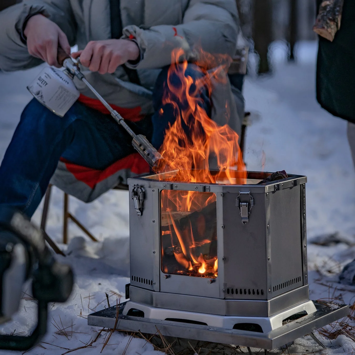 雪が積もった屋外で、一人の人物がポータブルな焚き火台にバーナーを使って火を起こしている様子