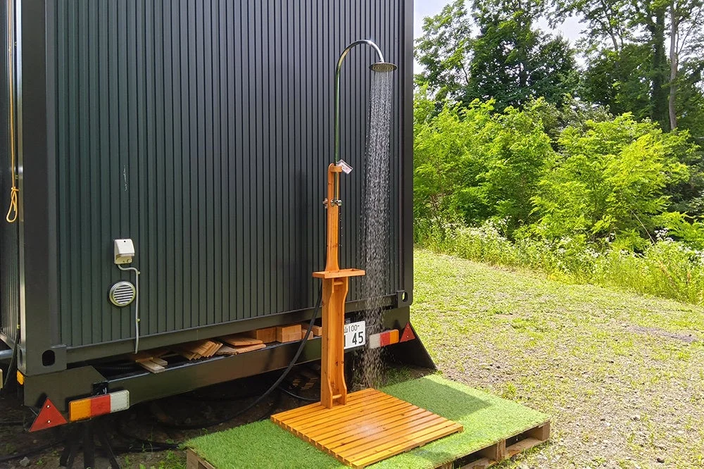 森を背景にした屋外に設置されたシャワーの様子。黒いコンテナのような建物の脇に木製のシャワースタンドがあり、水が勢いよく出ています。人工芝とウッドデッキが足元に敷かれています。