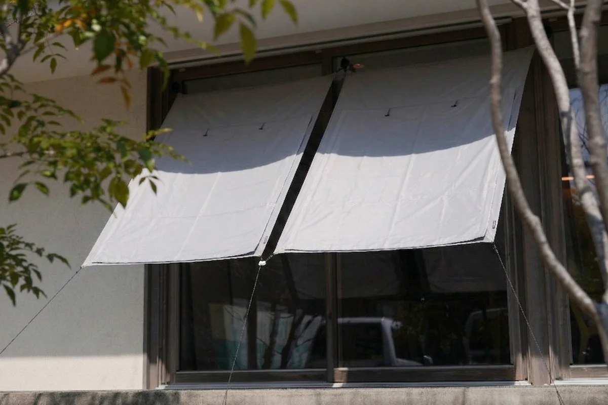 建物の窓に設置されたグレーの日よけが特徴的な画像です。窓の外には緑豊かな葉が茂っており、自然と建築が調和した穏やかな雰囲気を醸し出しています。