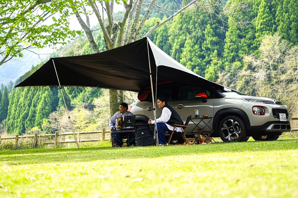 新緑の季節、2人の男性がSUVに連結したタープの下でくつろいでいる様子。自然豊かな場所で、キャンプやアウトドアを楽しんでいる光景です。