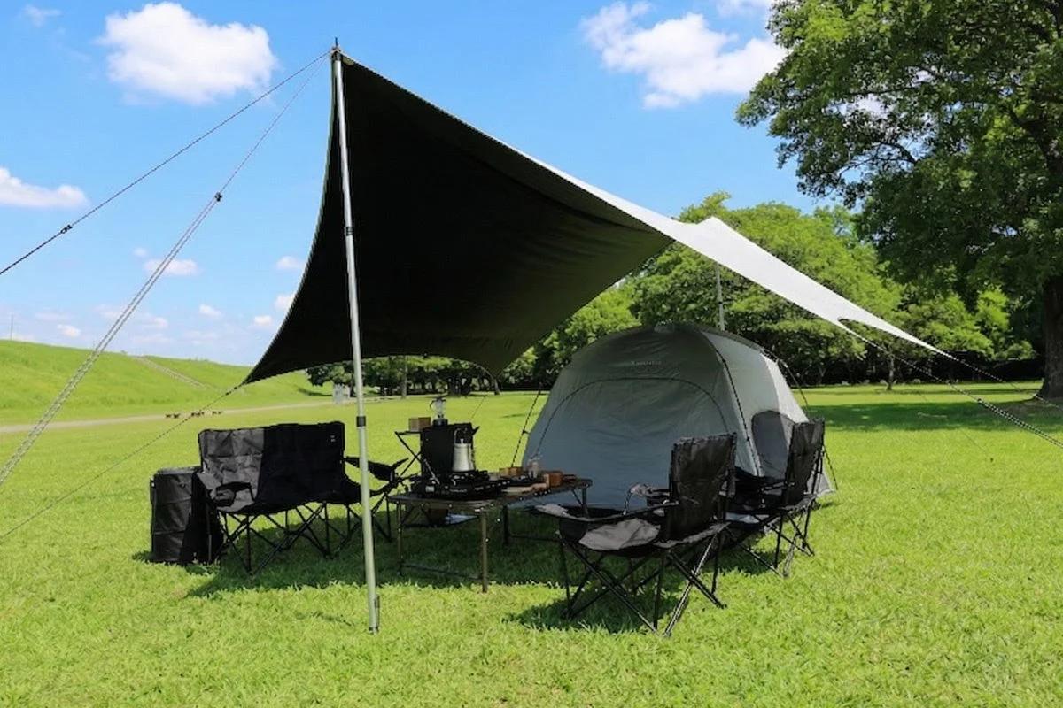 青空の下、広々とした芝生でキャンプを楽しんでいる様子。タープとテントが設置され、テーブルと椅子が並べられている。