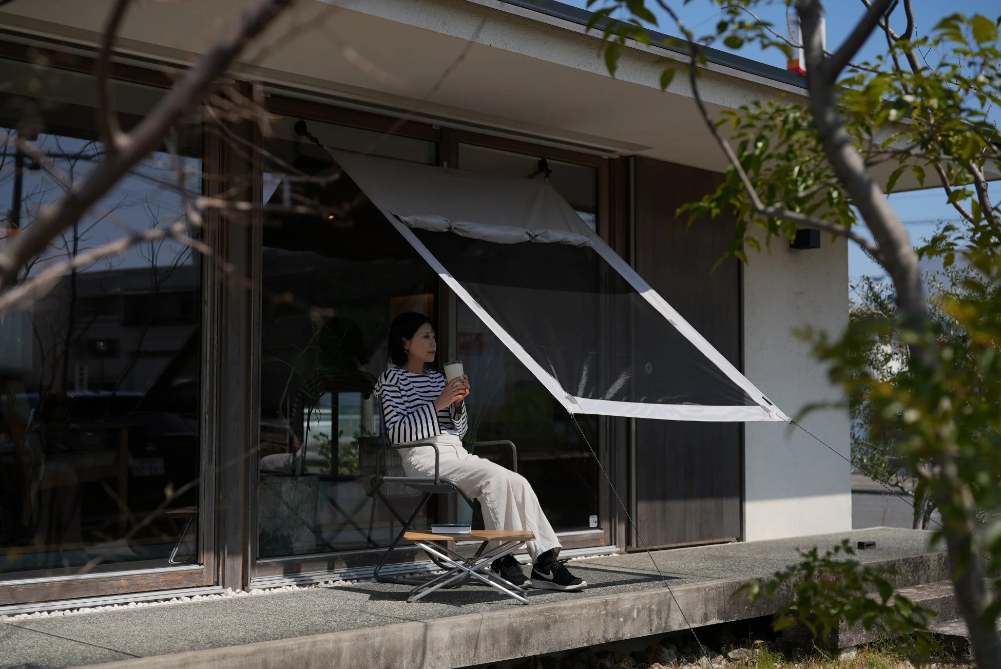 現代的な家屋のテラスで、女性が折りたたみ椅子に座り、マグカップを片手にくつろいでいます。日よけのタープが設置され、屋外でリラックスした時間を過ごしている様子です。