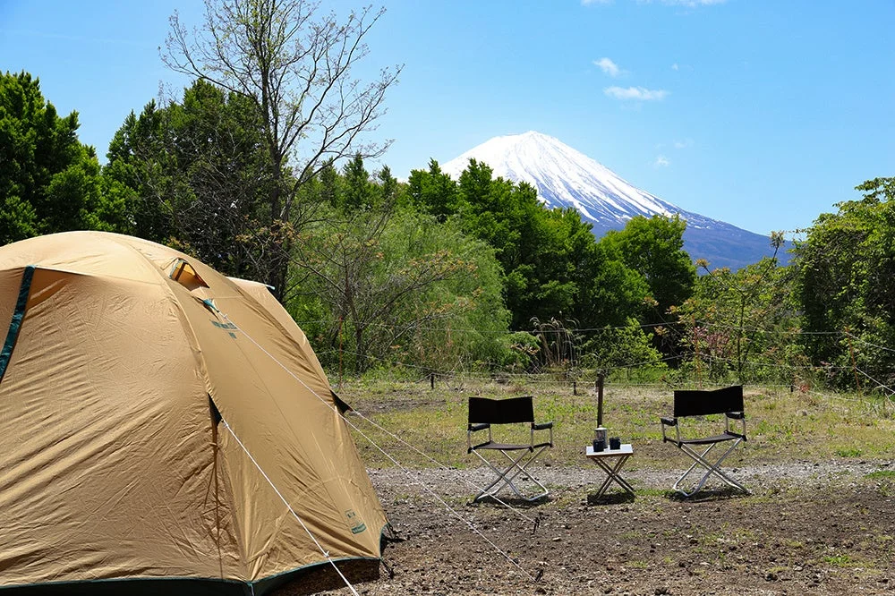 富士山を望むキャンプ場で、茶色のテントが設営され、折りたたみ椅子とテーブルが置かれています。新緑が美しい季節の晴れた日、アウトドアを楽しむ様子を捉えた一枚です。