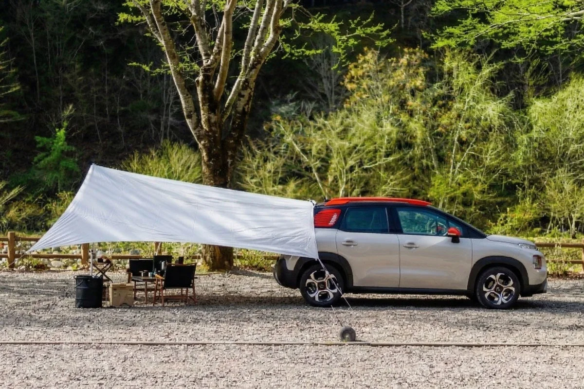 森を背景にした砂利の広場で、ベージュ色のSUVが白いタープと連結されています。タープの下にはキャンプ用の椅子やテーブルが置かれ、自然の中でのんびりとしたアウトドアシーンが広がっています。