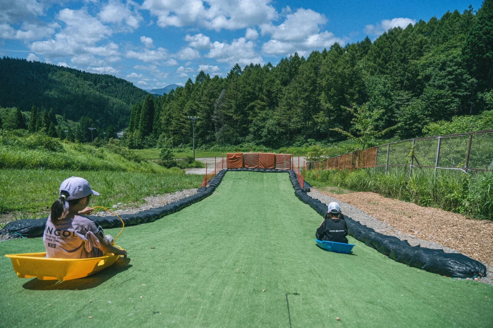 晴れた日に、二人の子供が人工芝のソリゲレンデでソリ遊びを楽しんでいる様子